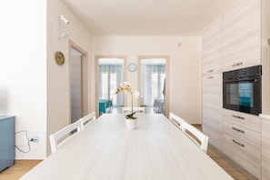 Uma cozinha moderna com forno embutido, um espaço de refeições com mesa e cadeiras brancas, e uma orquídea em vaso sobre a mesa.