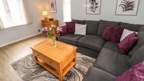 Sala de estar com sofá cinzento, almofadas roxas, mesa de centro de madeira e um vaso com flores.