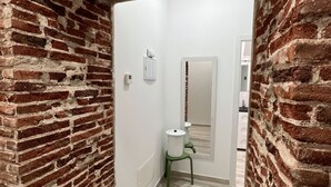 A narrow hallway with exposed brick walls and a light wooden floor. A small green stool with a white container is placed against the wall.