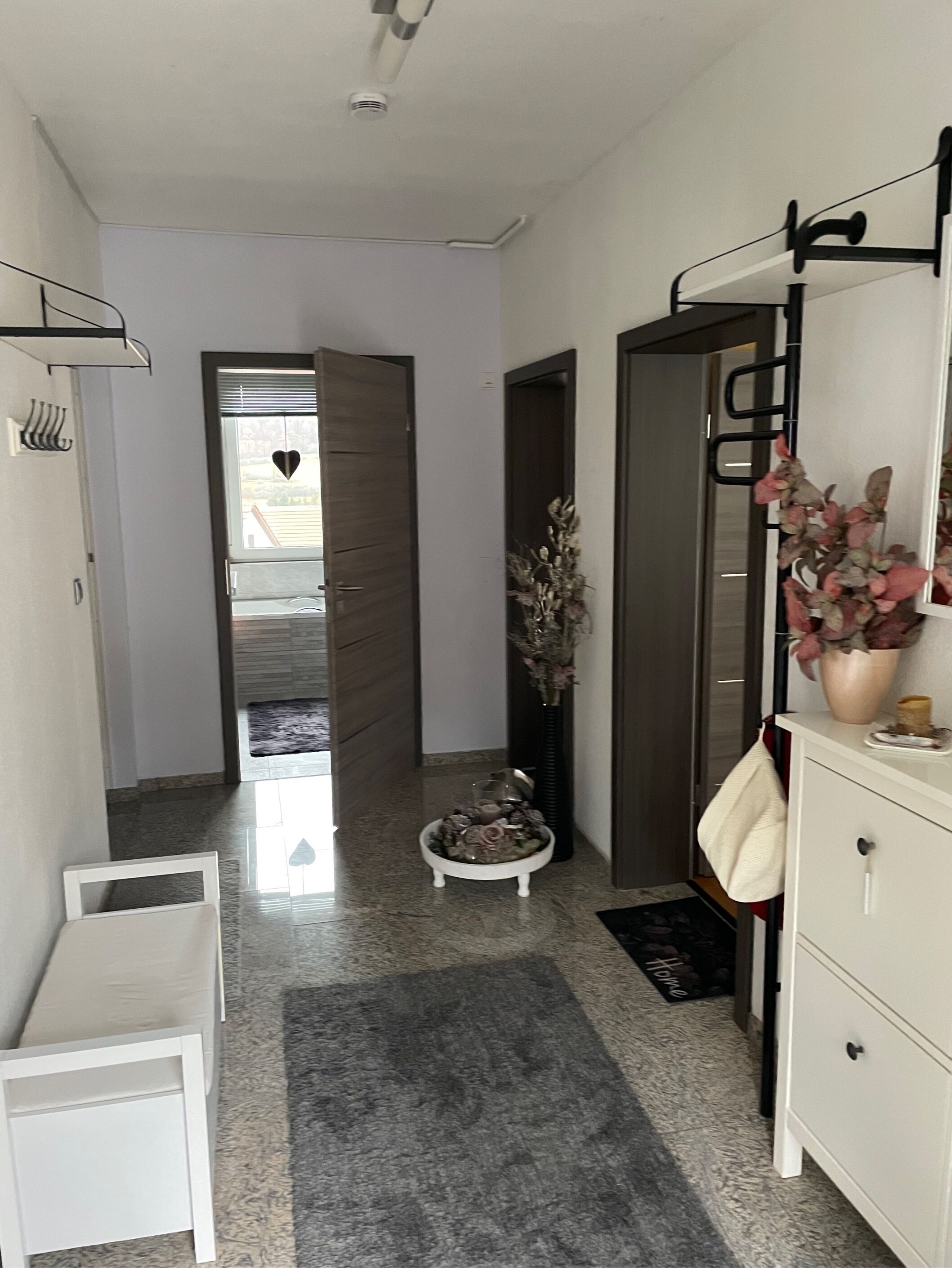 A hallway with a white bench, a small table with decorative items, a vase with flowers, and a white cabinet with a mirror.
