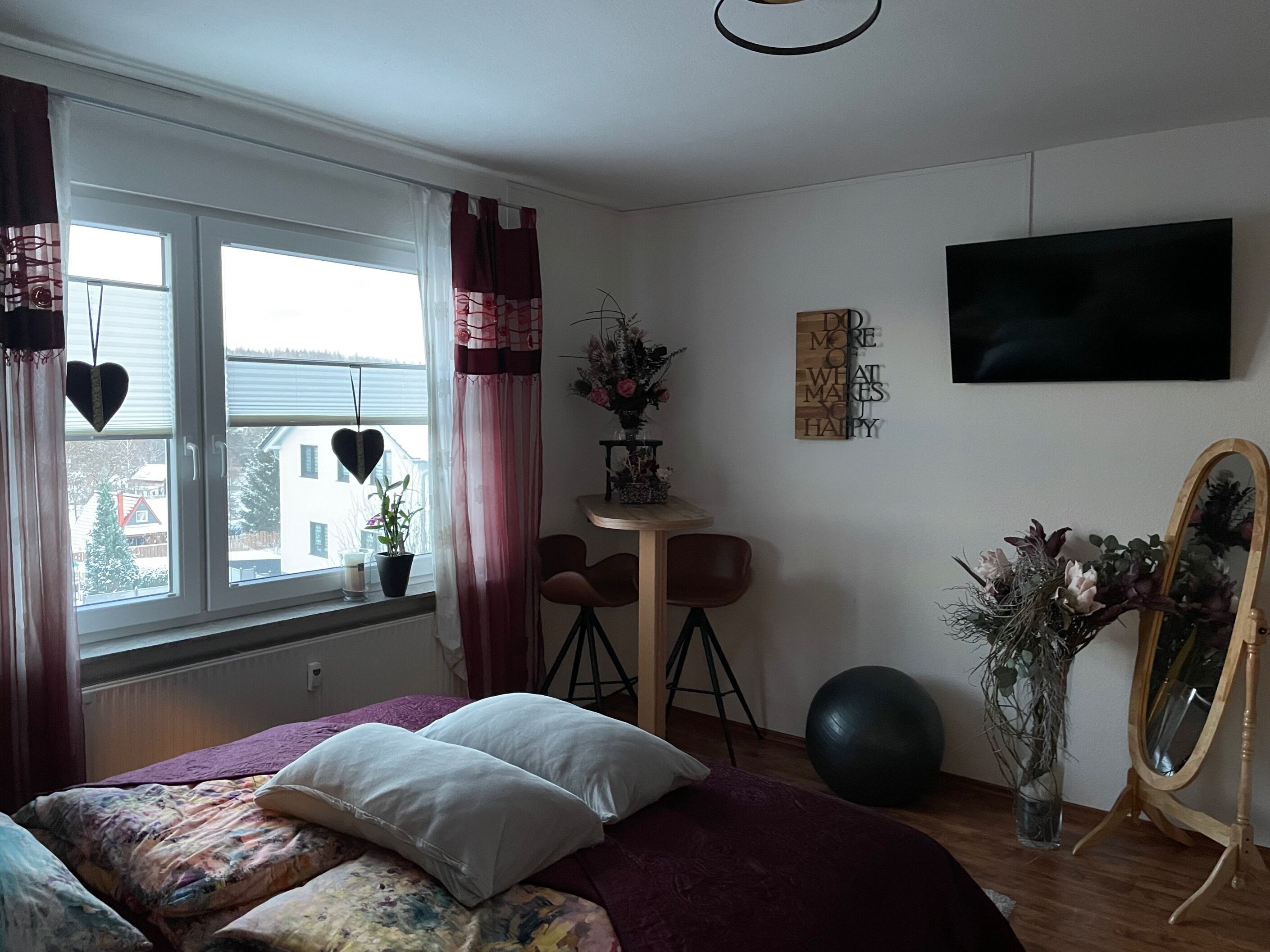 A bedroom with a bed, a window with curtains, a desk with a chair, a mirror, and a wall-mounted TV.