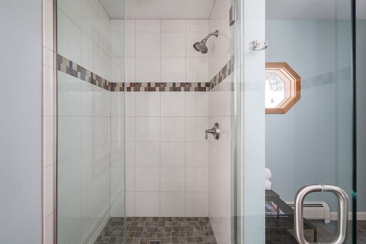 A glass-enclosed shower with white tiles and a brown-tiled floor, a towel rack, and a small window with a geometric design.