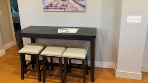 Een eetgedeelte met een donker houten tafel en drie bijpassende stoelen, een schilderij van een pier in de zonsondergang, en een wit oppervlak met structuur.