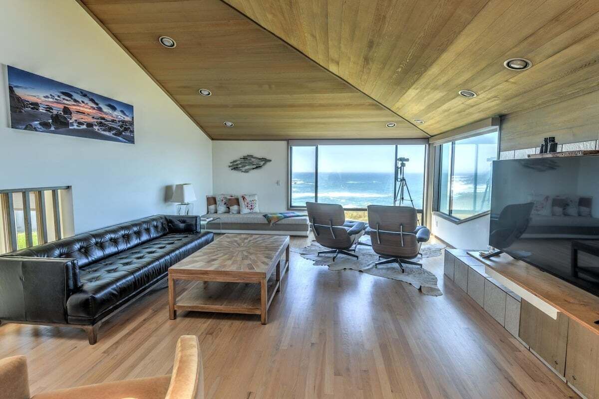A modern living room with a black leather sofa, wooden coffee table, and two armchairs. A large TV is mounted on the wall, and there is a window with a view of the ocean.