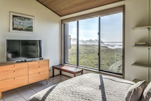 A bedroom with a large window, a bed, a TV on a wooden dresser, and a bench. - Sea Break | Coastal Escape with Hot Tub (Sea Ranch)