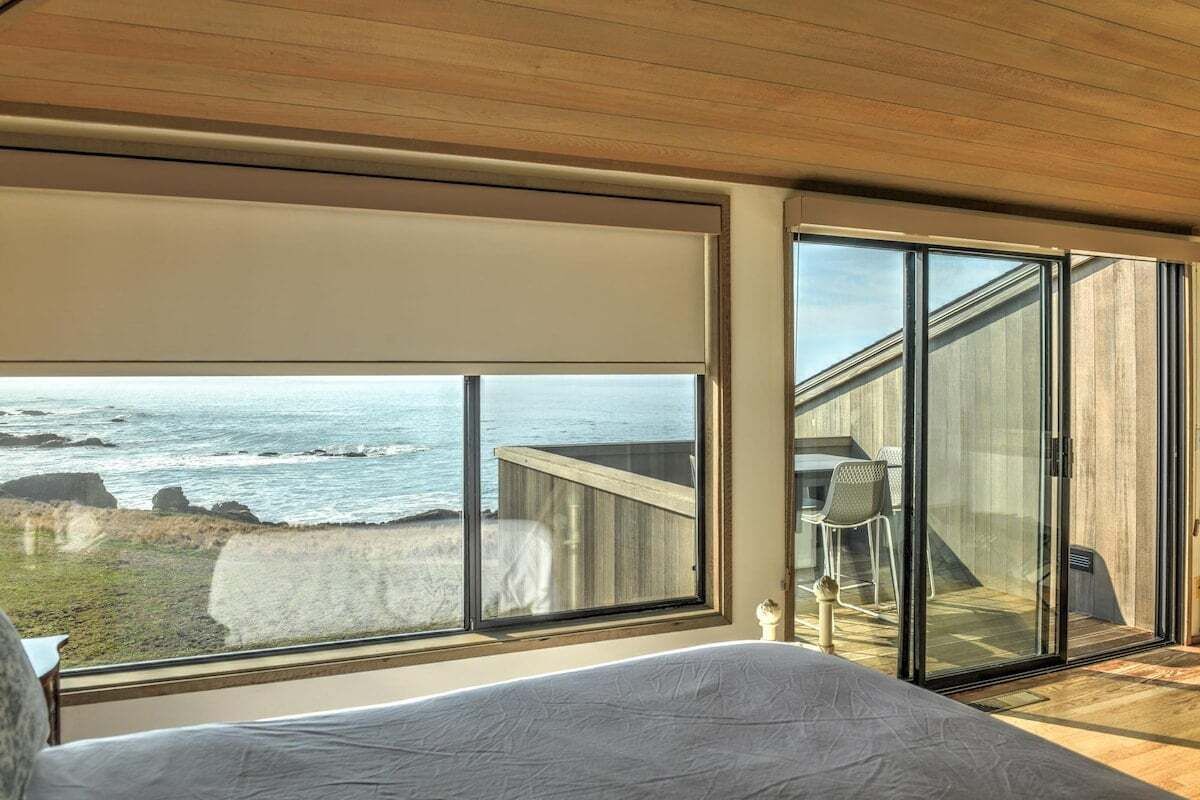 A bedroom with a large window overlooking the ocean, a bed, and a sliding glass door leading to a balcony.