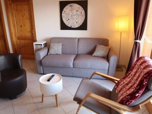 Uma sala de estar com um sofá cinzento, uma poltrona preta, uma mesinha lateral branca e um cobertor vermelho estampado.