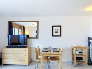 A modern room with a flat-screen TV on a wooden cabinet, a dining table set with plates and glasses, a small framed picture on the wall, and a coffee machine.