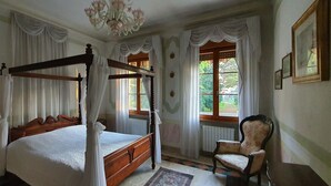 A four-poster bed with a canopy, a chair, and a chandelier in a room with large windows and curtains.