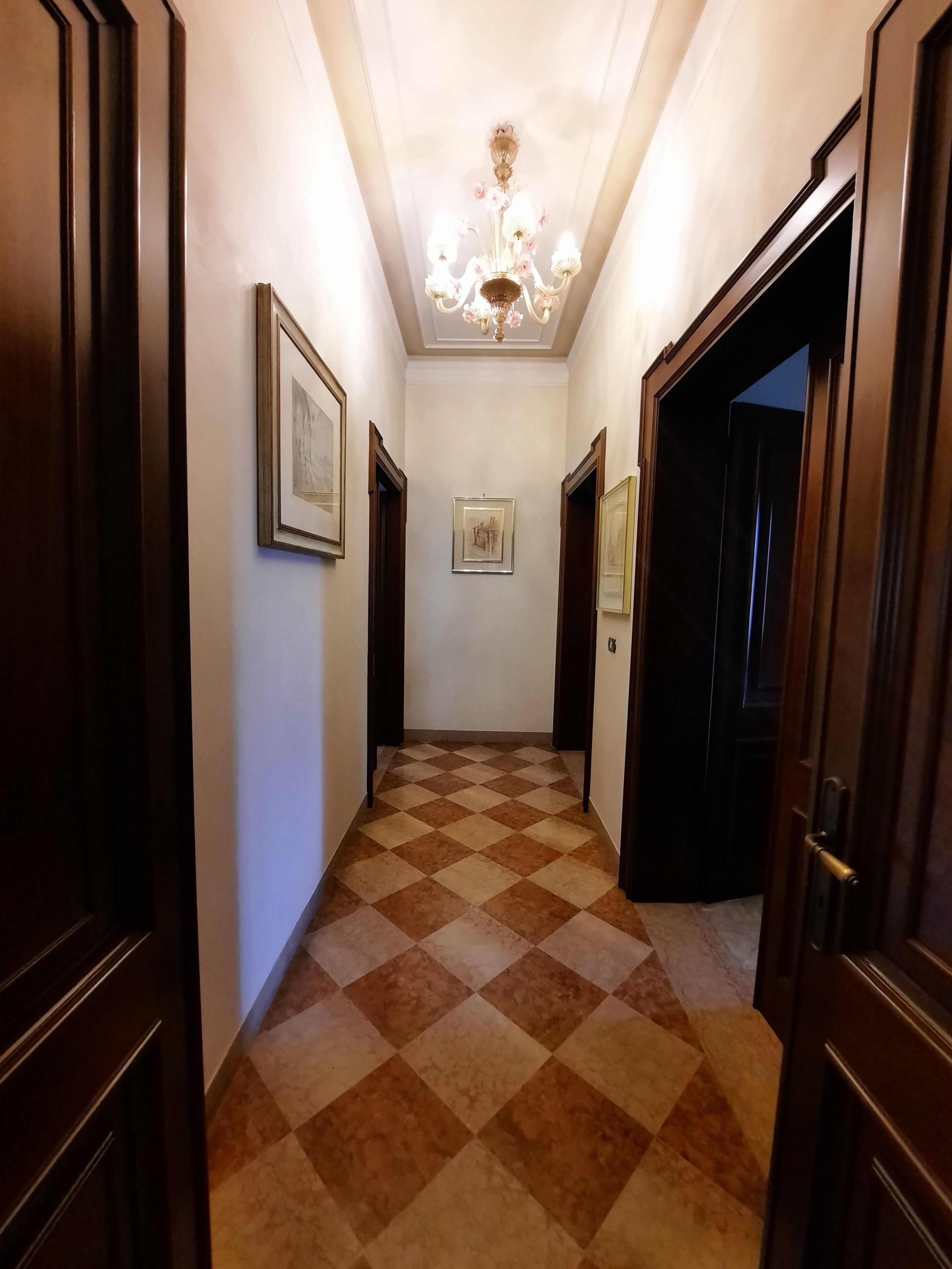 A hallway with a checkered floor, framed artwork on the walls, and a chandelier.