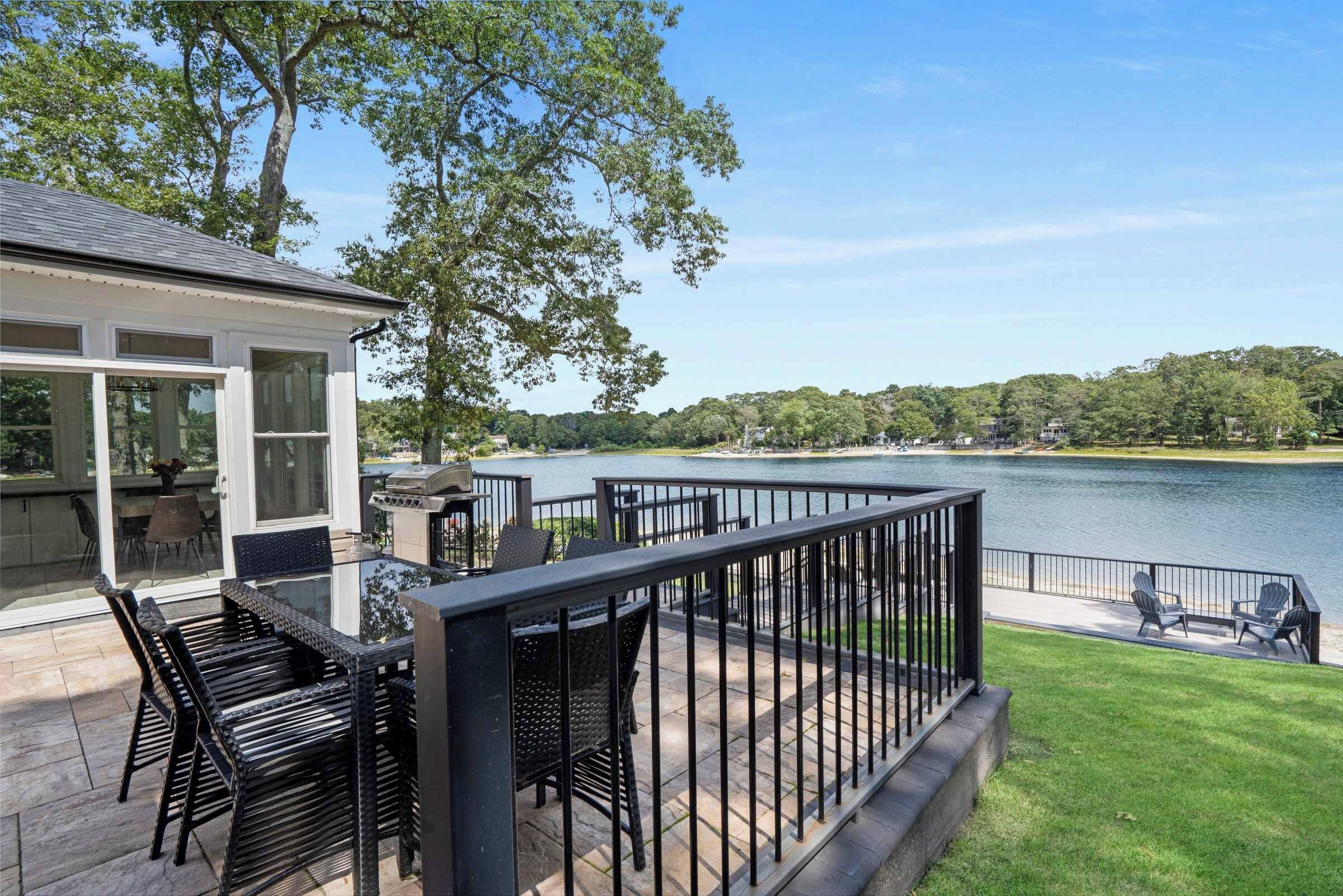 A deck with outdoor furniture overlooking a lake, a barbecue grill, and a view of a lake and trees.