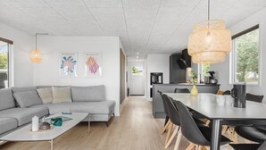 A modern living space with a grey sofa, a white coffee table, and a dining area with black chairs and a wooden table. There are two wall-mounted artworks, a kitchen with a black oven and a coffee machine, and a large window providing natural light. - Awesome home in ørsted with WiFi (Ørsted)