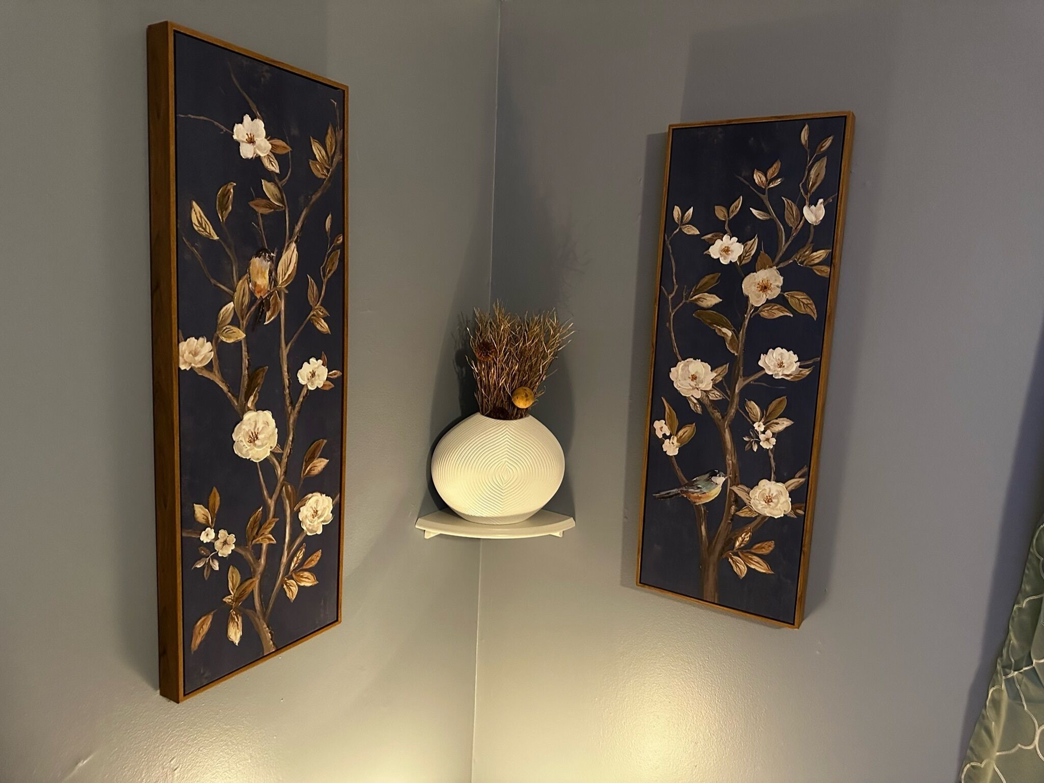 Deux tableaux accrochés au mur, ornés de motifs floraux et d’oiseaux, un vase blanc contenant des plantes séchées et, entre les deux, une étagère blanche.