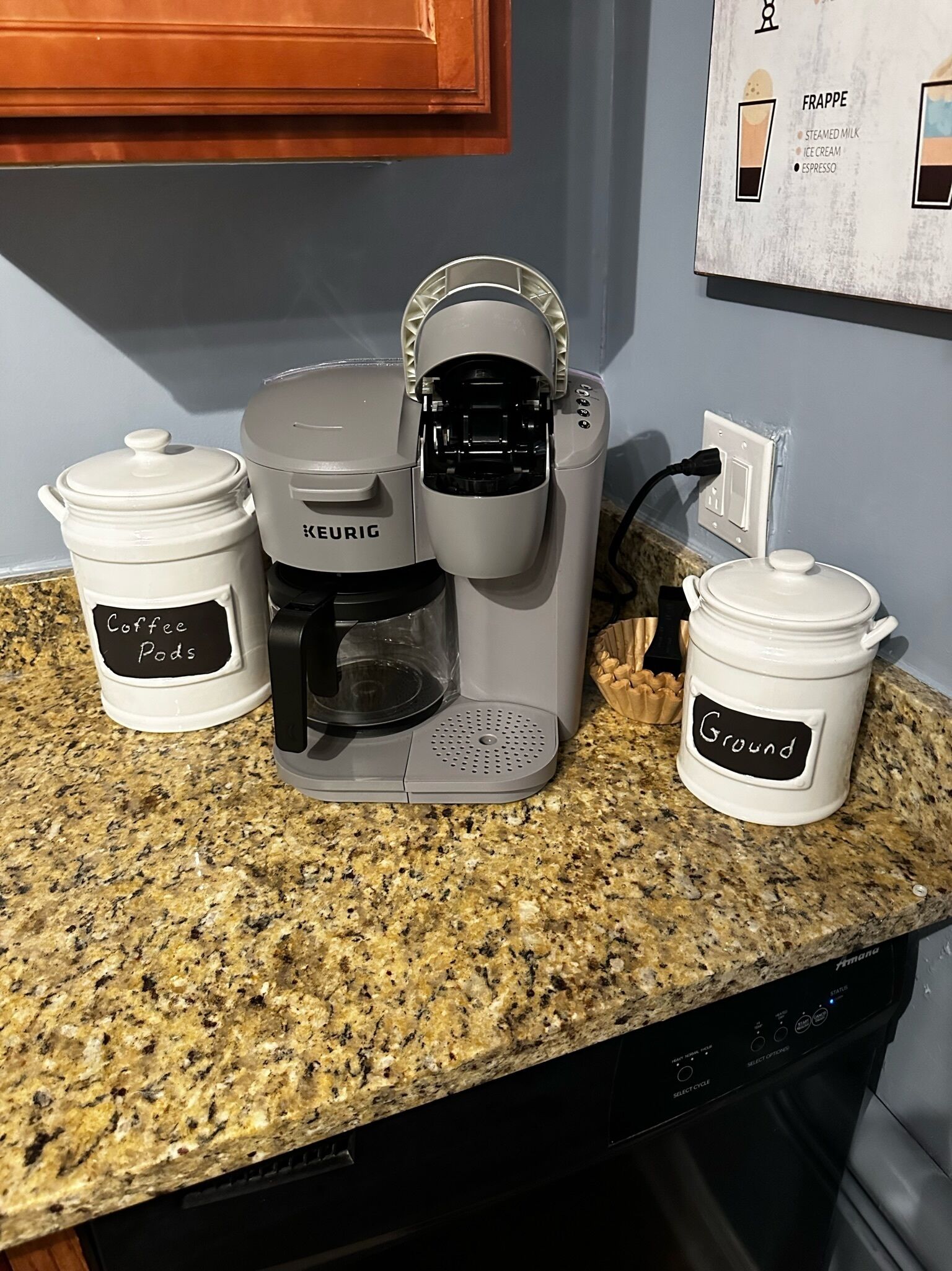 Una cafetera Keurig sobre una encimera de granito con dos cápsulas de café identificadas, una que dice 'Cápsulas de café' y la otra 'Café molido'.