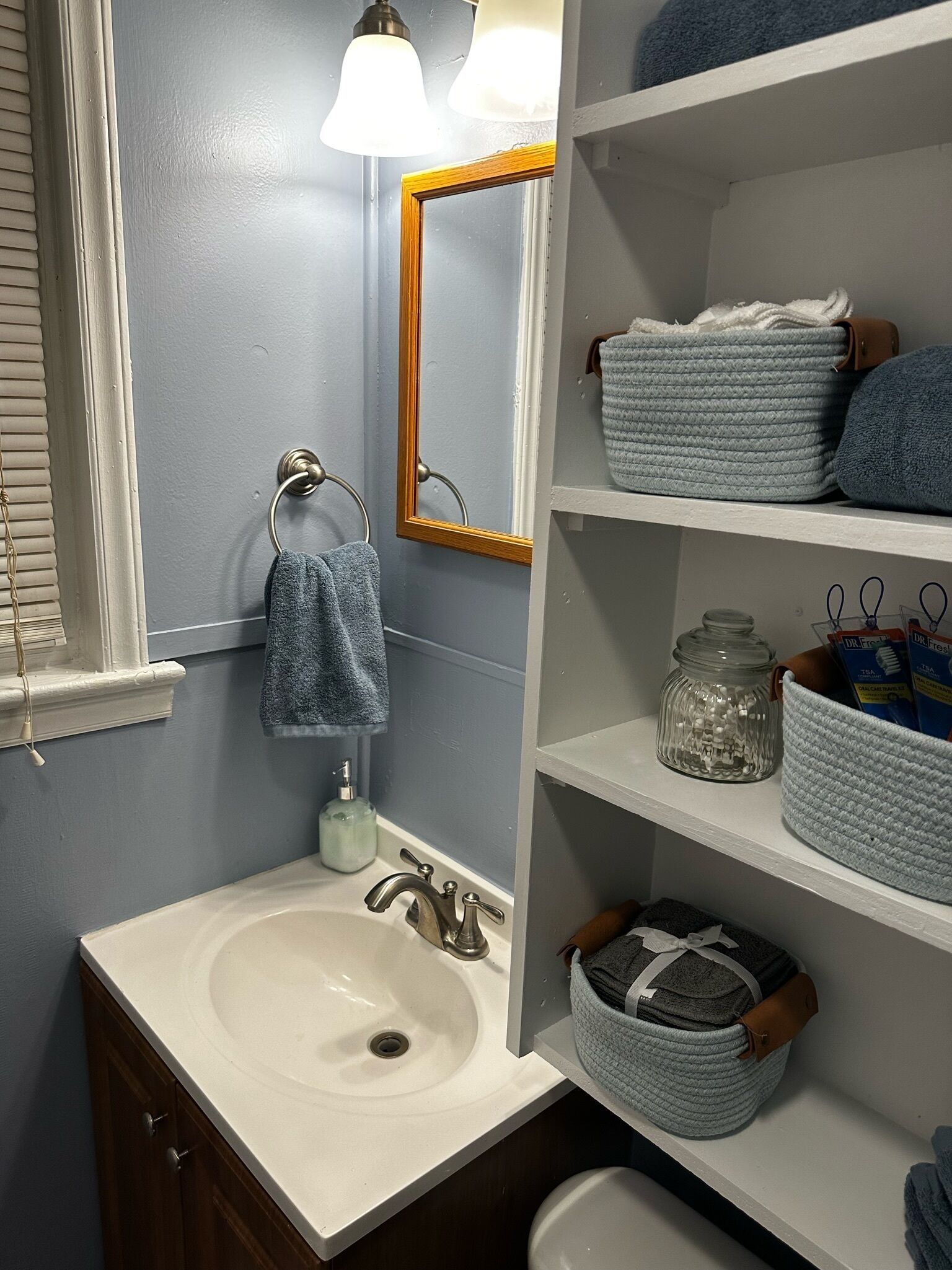 A bathroom with a white sink, a mirror, a towel rack, shelves with towels and toiletries.