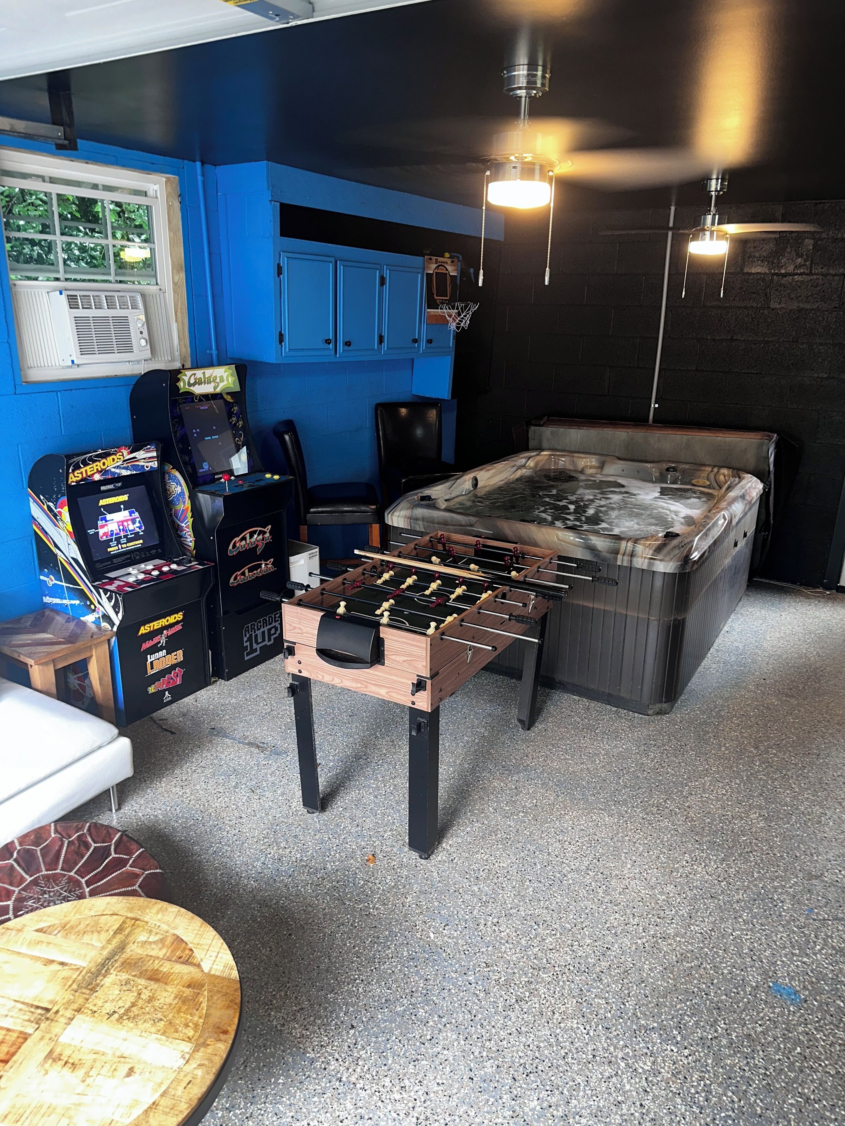 A room with a foosball table, two arcade games, a hot tub, and a window with blinds.