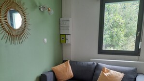 Un salón con un sofá gris, dos cojines estampados, un taburete de madera redondo, una alfombra tejida, un espejo montado en la pared y una ventana con vistas a zonas verdes.