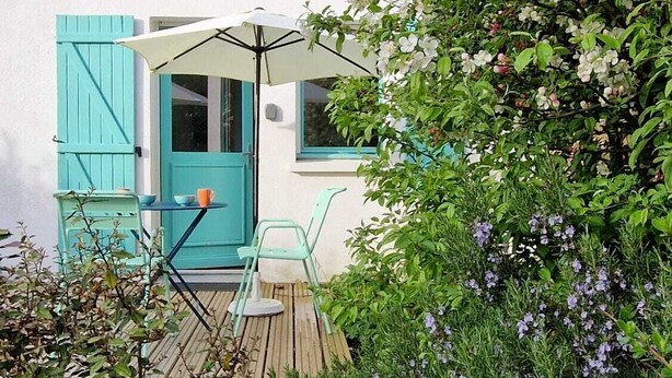 Una casa encantadora con tejado de tejas, un pequeño balcón con mesa y sillas, y un jardín con flores moradas.