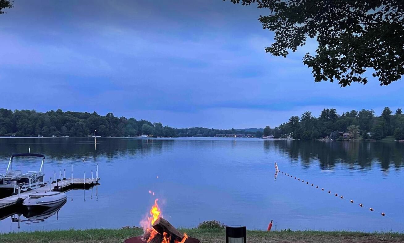 A calm lake with a dock and a campfire, surrounded by trees.