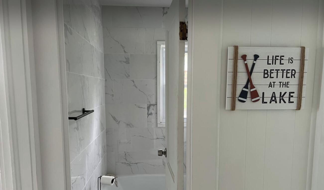 A bathroom with a shower, a toilet, and a towel rack. There is a sign on the wall with a message about life being better at the lake.