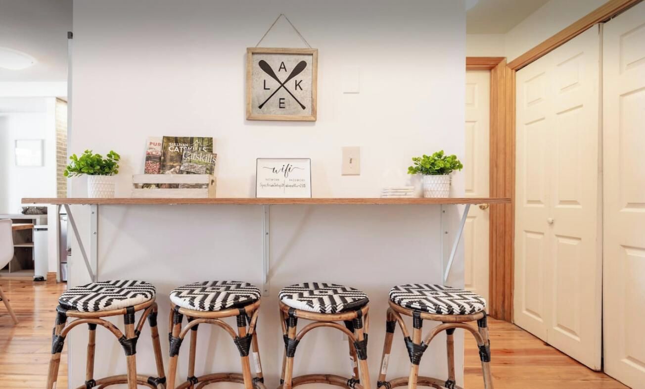 Una habitación con pared blanca, estante de madera con libros y un cartel enmarcado, tres taburetes de mimbre y una puerta.