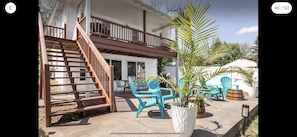 Une maison avec un escalier en bois menant à un balcon, un espace extérieur aménagé avec des chaises turquoise et une grande plante en pot.
