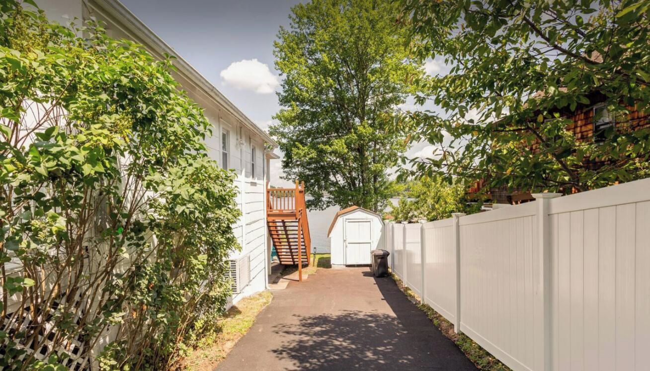 A narrow pathway leading to a white garage door, flanked by a white fence and dense foliage.