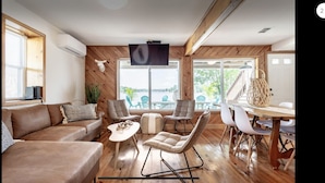 A spacious living room with a wooden floor, a brown leather sofa, a white coffee table, and a dining area with a wooden table and white chairs.
