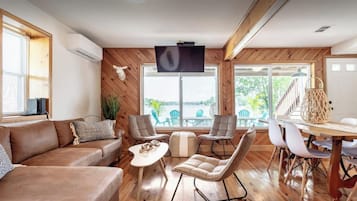 Un salón amplio con piso de madera, un sofá de cuero marrón, una mesita de café blanca y un comedor con mesa de madera y sillas blancas.