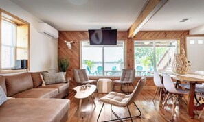 A spacious living room with a wooden floor, a brown leather sofa, a white coffee table, and a dining area with a wooden table and white chairs.