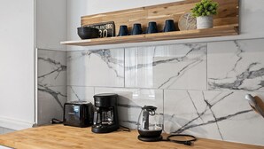A kitchen with a wooden shelf holding mugs and a small potted plant, a coffee maker, a toaster, and a kettle on a marble-tiled backsplash. - Center of Little Italy #4 (Cleveland)
