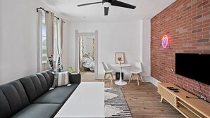 A modern living room with a black ceiling fan, a brick accent wall, a flat-screen TV, a wooden TV stand, a white coffee table, a grey sofa, and a small white table with chairs. - Center of Little Italy #4 (Cleveland)