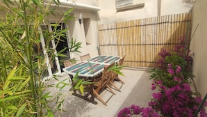 Un patio con mesa y sillas de madera, rodeado de plantas de bambú y flores moradas.