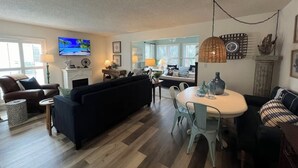 A spacious living room with a fireplace, a dining area with a round table and chairs, and a kitchen in the background. - Boutique Beachside condo with easy access to beach (St. Augustine)