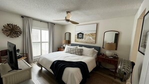 A bedroom with a bed, a desk, a chair, a ceiling fan, a window with curtains, and artwork on the walls. - Boutique Beachside condo with easy access to beach (St. Augustine)