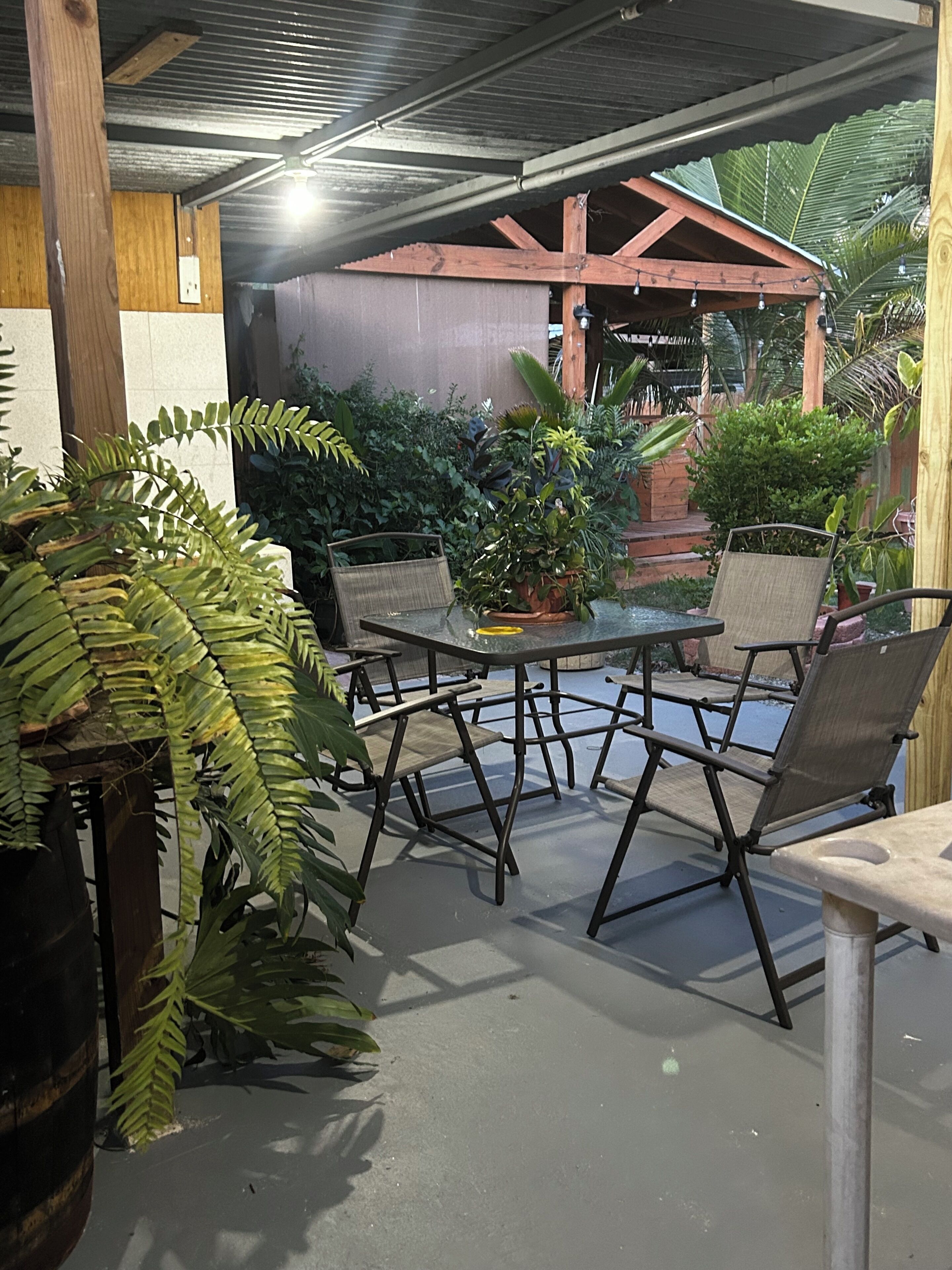 Un espace patio extérieur avec une table ronde et quatre chaises, entouré de différents pots de plantes et d’une structure de pergola en bois.