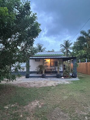 A small house with a green roof, surrounded by trees and a palm tree, with a gravel path leading to the entrance.