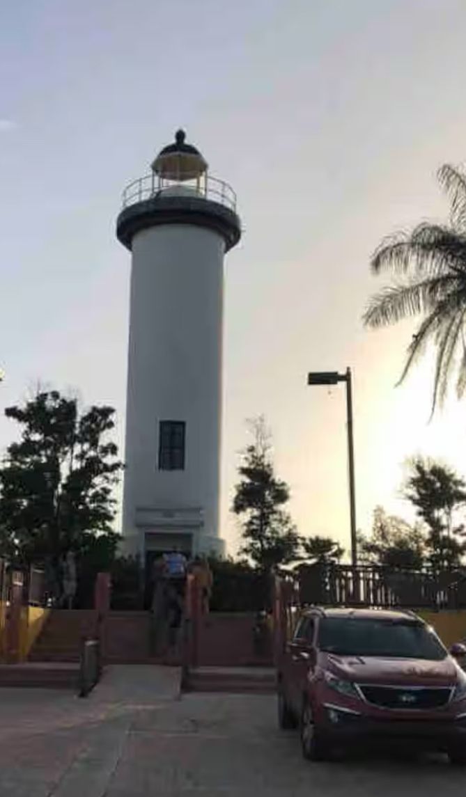 Un grand phare cylindrique, doté d’un balcon près du sommet, entouré d’arbres et bordé d’une zone pavée où est garée une voiture.