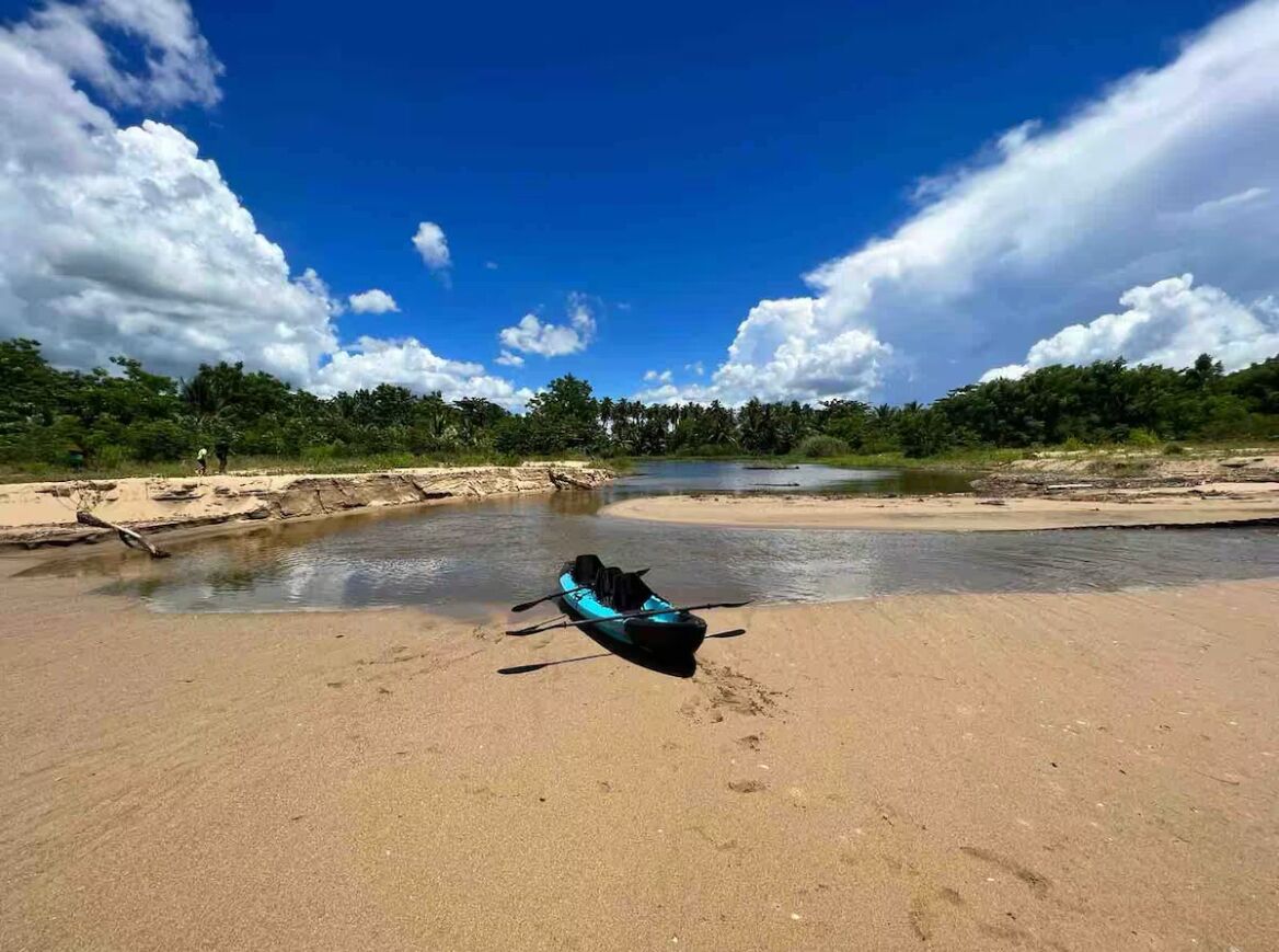 Een kajak op een zandstrand, met een rivier en bomen op de achtergrond.