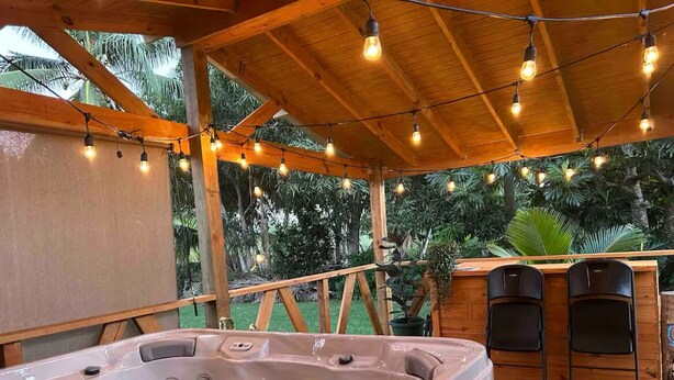Un bain à remous installé sous une pergola en bois, illuminé de guirlandes lumineuses, et entouré de verdure.