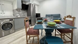 Ett modernt kök med rostfria vitvaror, ett matbord dukat med färgglada tallrikar och glas, och en soffa i bakgrunden.