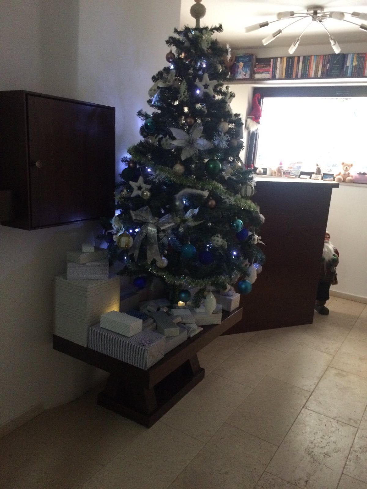 A Christmas tree with ornaments and lights, placed next to a dark wooden cabinet and a small table with wrapped gifts.