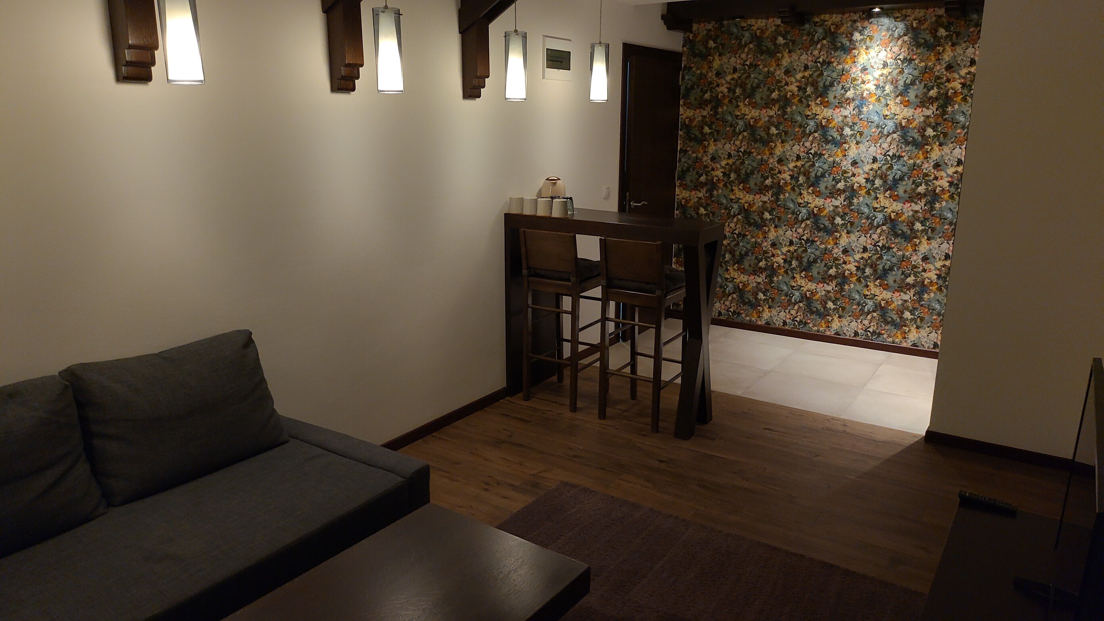 Una sala moderna con un sofá oscuro, una mesa de centro de madera, y un comedor con dos sillas y una pequeña mesa. Hay lámparas colgantes sobre el sofá y un papel tapiz floral en la pared.