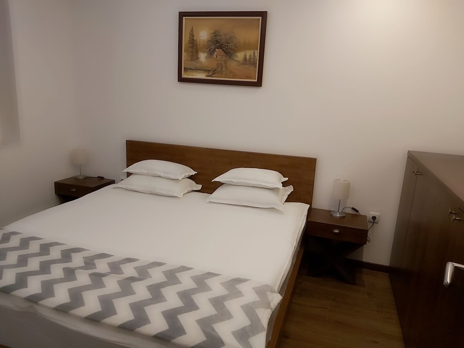 A neatly made bed with a patterned bedspread, a wooden headboard, two bedside tables with lamps, and a painting on the wall.