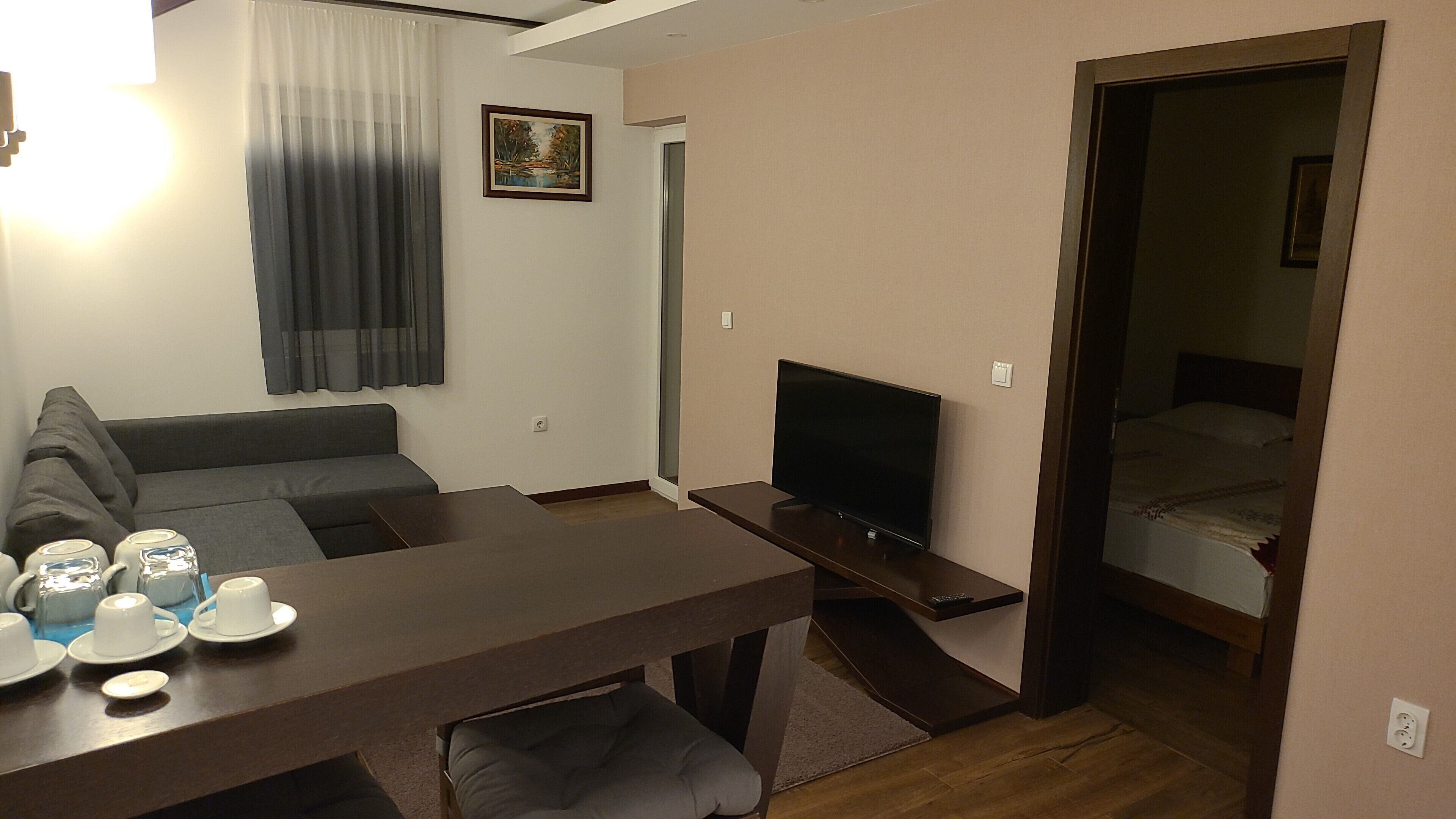 Een knus woongedeelte met een bank, een tv op een meubel, een eettafel met kopjes en schotels, en een deur naar een slaapkamer.