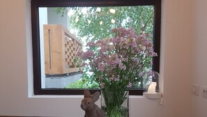 Una ventana con vista a un edificio y árboles, un florero con flores y una pequeña figurina sobre una superficie oscura.