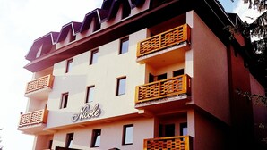 Un hotel de varios pisos con balcones, un letrero que dice 'Nicole' y mobiliario exterior con sombrillas.