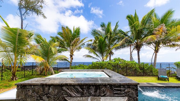 A stone hot tub with a view of the ocean, surrounded by palm trees and a fence.