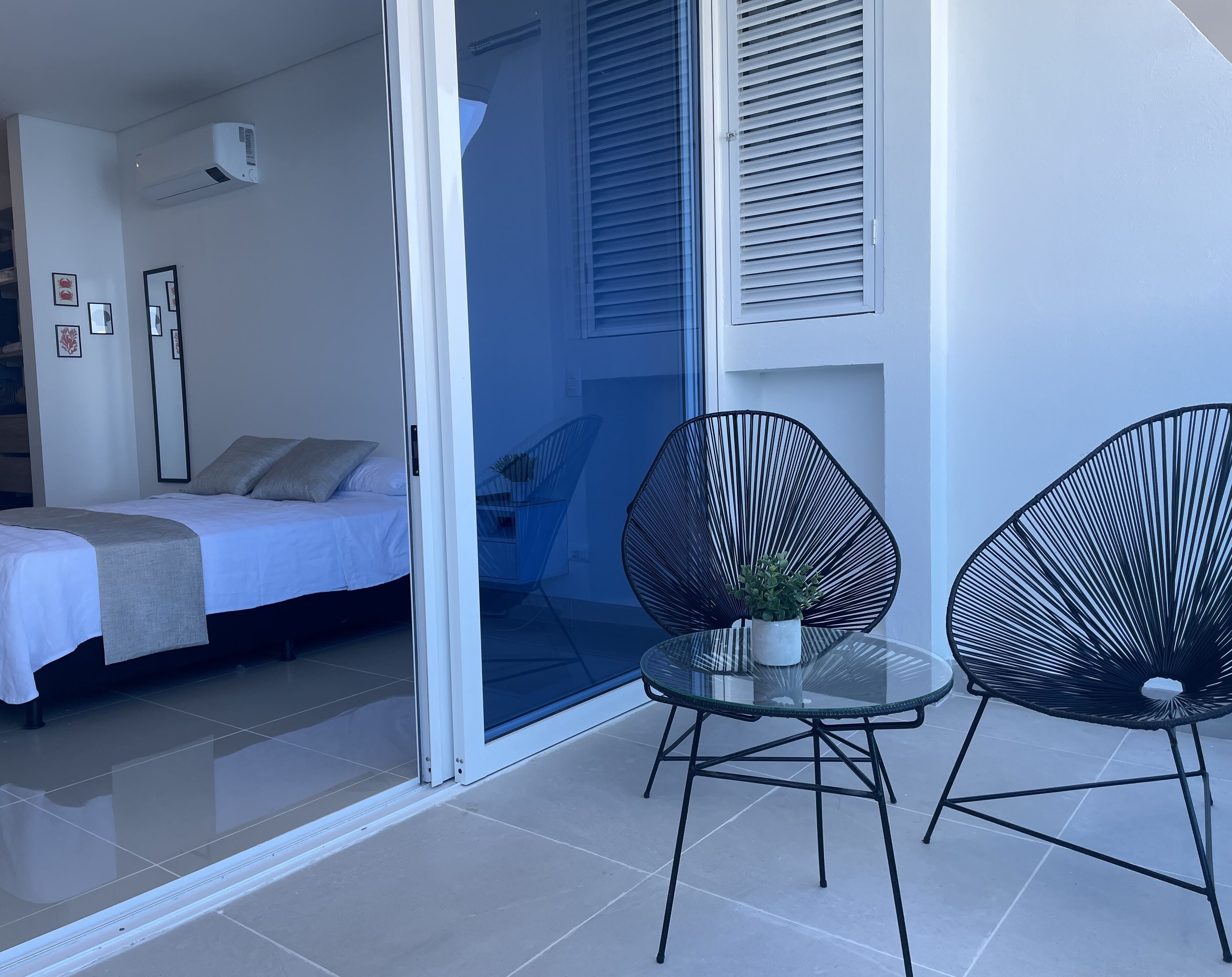 Une chambre moderne avec un lit, une porte vitrée donnant sur un balcon, et deux chaises noires accompagnées d’une petite table et d’une plante.