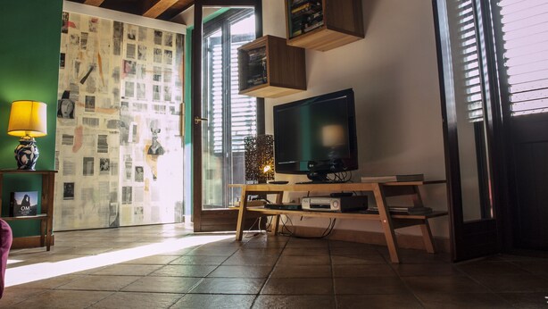 A living room with a television on a wooden stand, a green wall with newspaper clippings, and a window with blinds.